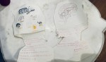 Two pieces of paper cut into head shapes lie on a table, each with drawings and handwritten notes. The left head has doodles of a house, sun, and smiley faces; the right head has a detailed drawing of a brain with red text around it.