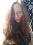 A woman with long, wavy brown hair and glasses smiles slightly. She wears a dark top, revealing a tattoo on her arm. The background features floral patterns with light streaming in, subtly symbolizing life's complexity akin to genetic mutations.