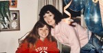 A woman in a pink sweater leans over a smiling young girl in a red Stanford sweatshirt. Amid the festive pattern of blue balloons and orange streamers, time seems to pause. A plant and photos subtly frame this heartfelt indoor scene.