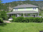 A two-story green house with a wraparound porch, perfect for observation of the surrounding forested hills, features lush shrubs and a well-kept lawn, offering both beauty and a sense of safety beneath the blue sky.