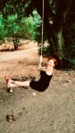 A woman with red hair, wearing a black dress and red shoes, smiles while swinging on a rope swing. It is hanging from a tree in a wooded outdoor area.