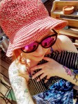 A person wearing a large red sun hat and sunglasses smiles while holding a striped towel, reminiscent of a vibrant Brass Monkey. They have colorful tattoos on their arms and are enjoying the great outdoors on a sunny day.