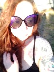 A person with long red hair and large, dark sunglasses stands under the bright sunlight. Their floral tattoos peek from beneath a black top, evoking the freedom of instinct. Sunlight partially obscures their face, like memories in a spotless mind.