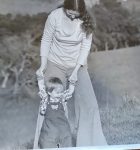 A woman holding the hands of a child who is learning to walk outside. The woman has long hair and is wearing a light-colored top and flared pants. The child is dressed in overalls and a patterned shirt. They are both smiling and enjoying a sunny day in a field. The background is slightly blurred with trees and nature.