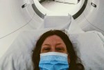 A person wearing a blue face mask lies on a hospital bed, about to enter a CT or MRI scanner. The circular opening of the scanning machine is visible in the background.