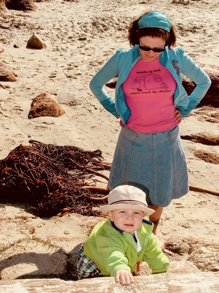A woman in a blue ja cket and pink shirt stands on a sandy beach, looking down at a smiling baby in a green jacket and hat, who is sitting and reaching up from the sand. Dried seaweed is visible on the beach.