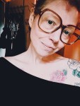 A person with large glasses and floral tattoos on their shoulder smiles slightly, wearing an off-the-shoulder black top. Warm lighting highlights their face and tattoos.