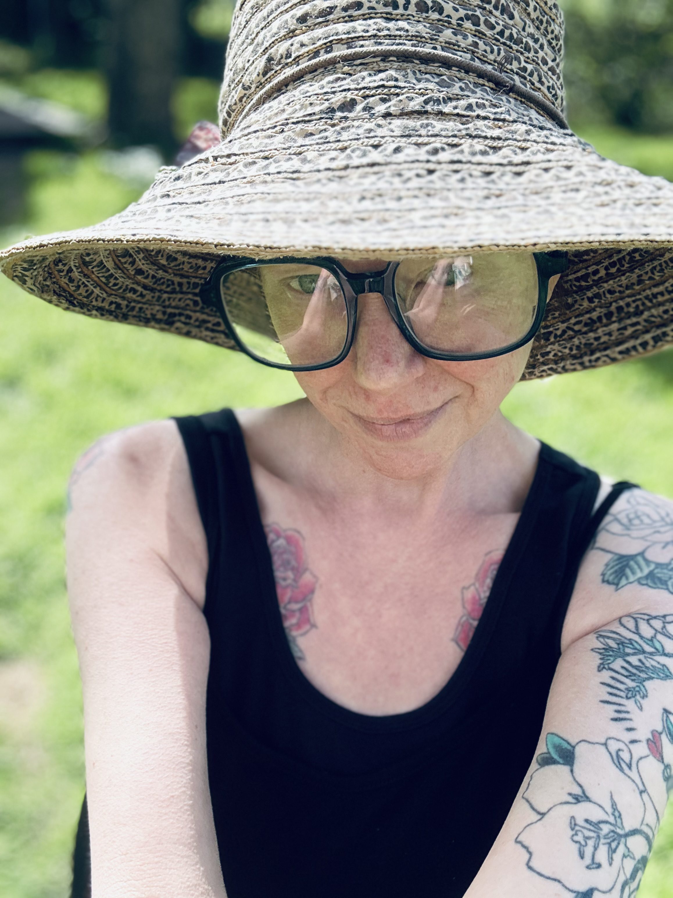 A person wearing a wide-brimmed hat, large glasses, and a sleeveless black top stands outside. They have floral tattoos on their chest and arm, and a soft smile. Greenery is blurred in the background.