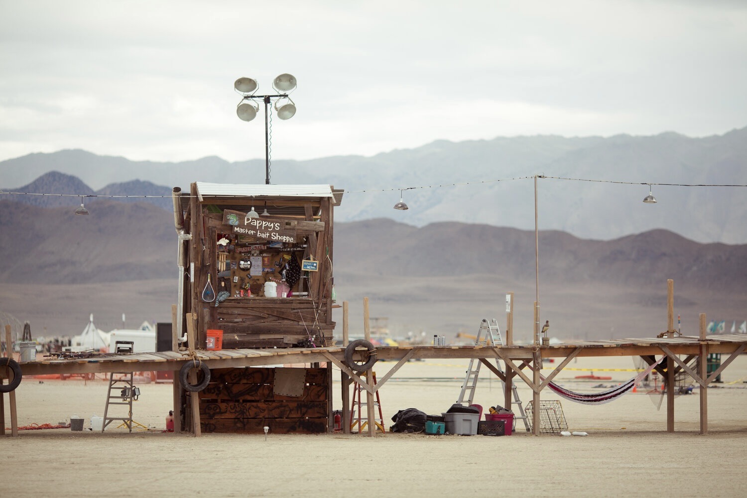 A small, rustic wooden shack labeled "Pappy's Blue Bottle Saloon" stands on a raised platform in a desert, with scattered chairs and items around it and mountains in the background.