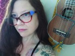 A woman with long brown hair and red glasses looks to the side. She has a floral tattoo on her shoulder and sits next to a wooden ukulele hanging on the wall by yellow yarn.