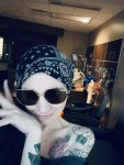 A woman wearing sunglasses and a black bandana with white patterns smiles and poses with her hand near her face. She has a large rose tattoo on her shoulder and sits enjoying the hospital view in a bright hospital room.